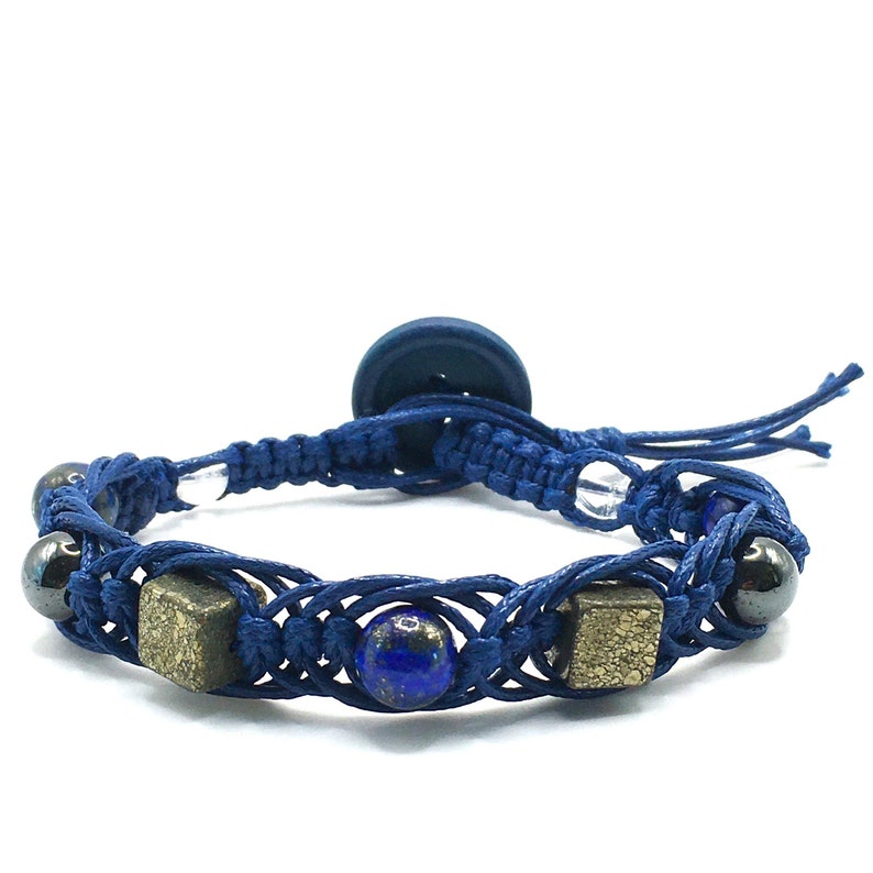 Pode incluir: Um bracelete de macram&eacute; azul com fecho de bot&atilde;o. O bracelete apresenta contas azuis e douradas, e um bot&atilde;o azul escuro.