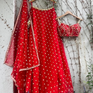 May include: A striking red Indian lehenga set, comprising a skirt, blouse, and dupatta, embellished with gold detailing. The skirt and dupatta have a polka dot design. The blouse is intricately embroidered. The set is displayed on wooden hangers.