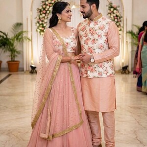 May include: A couple in formal attire, likely for a wedding. The woman wears a pink lehenga with gold trim and a matching dupatta. The man wears a peach kurta and pants with a floral vest. They stand in front of a floral arch.