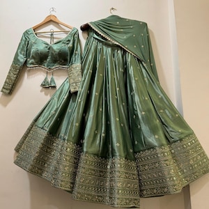 May include: A sage green Indian formal outfit, including a long-sleeved blouse with intricate embroidery on the sleeves and a matching skirt. The skirt features detailed gold embroidery along the hem. A matching scarf completes the ensemble.