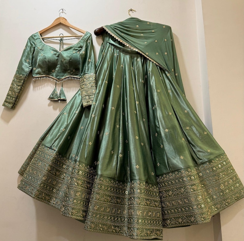 May include: A sage green Indian formal outfit, including a long-sleeved blouse with intricate embroidery on the sleeves and a matching skirt. The skirt features detailed gold embroidery along the hem. A matching scarf completes the ensemble.