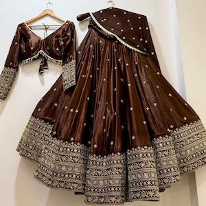 Puede incluir: Un conjunto de tres piezas de satén marrón, que incluye una blusa de manga larga, una falda amplia y una bufanda a juego. La falda y las mangas presentan intrincados bordados dorados. La blusa tiene cuello en V y cierre de lazo.