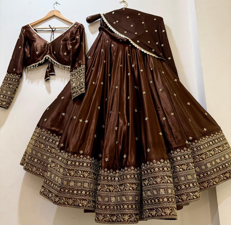 May include: A brown satin three-piece outfit, including a long-sleeved top, a full skirt, and a matching scarf. The skirt and sleeves feature intricate gold embroidery along the edges. The top has a V-neck and a tie closure.