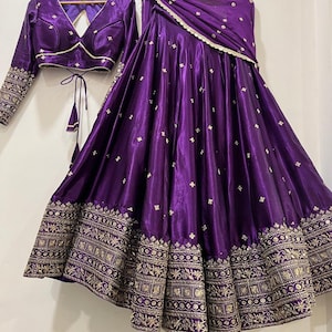 May include: A vibrant purple traditional Indian outfit, including a long skirt, a cropped top, and a matching scarf. The skirt and top feature intricate gold embroidery along the hems and sleeves, with delicate gold accents throughout the fabric.