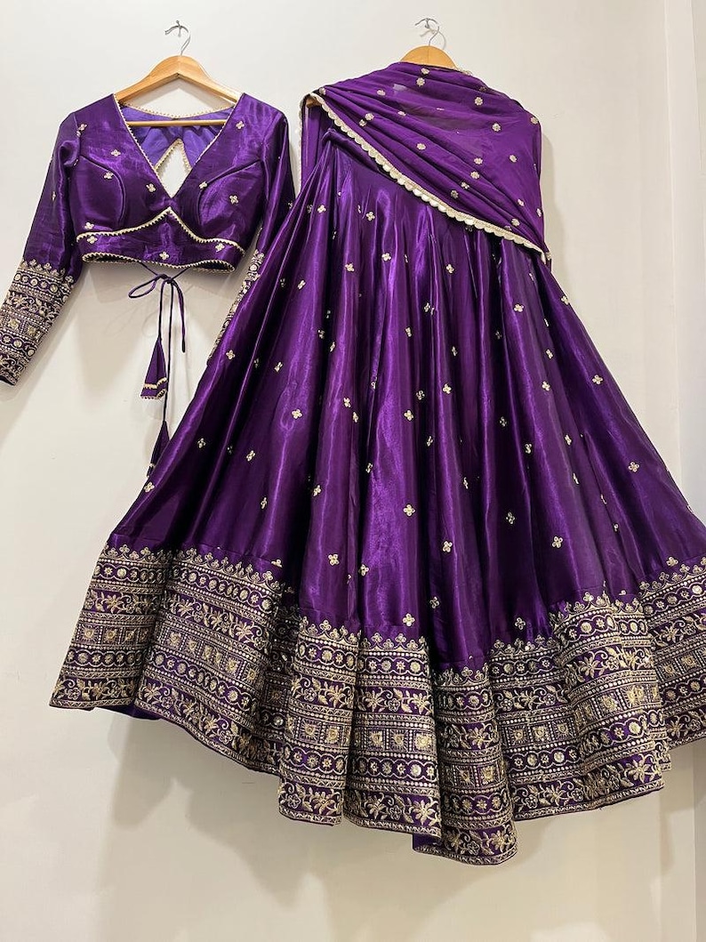 May include: A vibrant purple traditional Indian outfit, including a long skirt, a cropped top, and a matching scarf. The skirt and top feature intricate gold embroidery along the hems and sleeves, with delicate gold accents throughout the fabric.