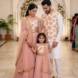 May include: A family dressed in matching traditional Indian attire. The women and girl wear pink skirts and tops with floral patterns, complemented by sheer scarves. The man wears a white outfit with a floral vest. The background features floral decorations.