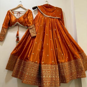 May include: An orange silk lehenga set, featuring a long skirt, a cropped blouse, and a matching scarf. The skirt and blouse have intricate gold embroidery. The blouse has long sleeves and a V-neckline. The set is displayed on a hanger.