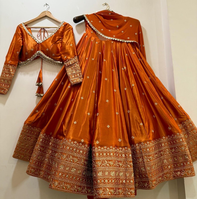 May include: An orange silk lehenga set, featuring a long skirt, a cropped blouse, and a matching scarf. The skirt and blouse have intricate gold embroidery. The blouse has long sleeves and a V-neckline. The set is displayed on a hanger.