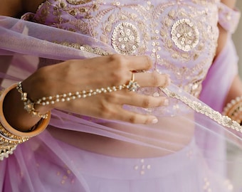 May include: A lavender-colored, embellished top and skirt set with gold accents. The top features intricate embroidery and sequins, while the skirt has scattered gold details. A sheer, matching lavender scarf drapes over the shoulders. Gold bangles and a hand chain complete the look.