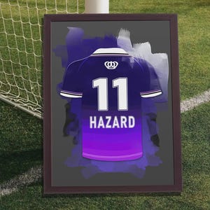 Puede incluir: Obra de arte enmarcada que presenta una camiseta de fútbol morada y blanca con el número 11 y el nombre HAZARD. El diseño de la camiseta se encuentra sobre un fondo gris oscuro con pinceladas moradas, enmarcado en un marco marrón oscuro. La obra se exhibe en un campo verde.