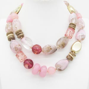 Pu&ograve; includere: Una collana a due fili con una variet&agrave; di perline nei toni del rosa, marrone e trasparente. Le perline includono forme ovali, rotonde e cilindriche, con dettagli color oro. La collana &egrave; esposta su una superficie bianca.