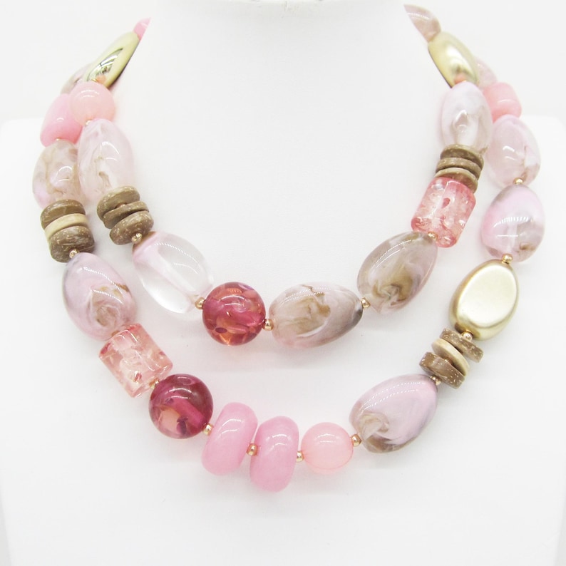 Pu&ograve; includere: Una collana a due fili con una variet&agrave; di perline nei toni del rosa, marrone e trasparente. Le perline includono forme ovali, rotonde e cilindriche, con dettagli color oro. La collana &egrave; esposta su una superficie bianca.
