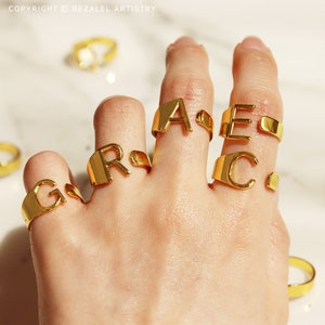 Op de afbeelding: Gouden ringen met de letters G, R, A, C en E erop. De ringen worden gedragen aan de vingers van een persoon.