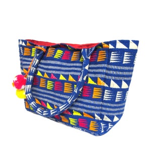 Może przedstawiać: Niebieska torba typu tote z kolorowym geometrycznym wzorem i ozdobnymi pomponami. Torba ma dwa uchwyty i czerwoną podszewkę.