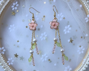 Op de afbeelding: Een paar delicate gouden oorbellen met roze bloemaccessoires en hangende parel- en groene bladeren.