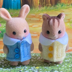以下が含まれることがあります： 2つのミニチュア動物フィギュア：ウサギと猫。ウサギは青いボタンが付いた水色のジャケットを着ており、猫は黄色のボタンが付いた黄色のジャケットを着ています。どちらも白い襟と黒い目です。
