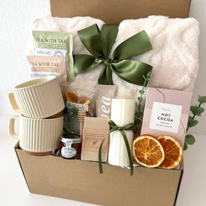 May include: A gift basket filled with self-care items. It includes a fluffy white blanket tied with a green ribbon, two cream-colored mugs, tea packets, a candle, hot cocoa, honey, soap, and dried orange slices. The box is labeled "Box 13".