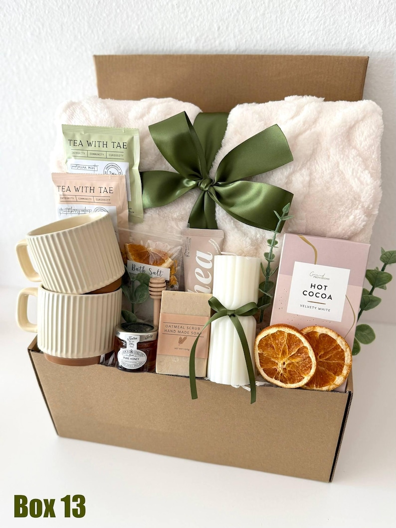 May include: A gift basket filled with self-care items. It includes a fluffy white blanket tied with a green ribbon, two cream-colored mugs, tea packets, a candle, hot cocoa, honey, soap, and dried orange slices. The box is labeled "Box 13".