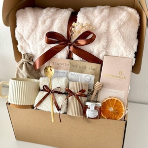 May include: A gift box filled with self-care items. It includes a fluffy white blanket, a mug, tea packets, a candle, honey, a wooden honey dipper, a bar of chocolate, and a dried orange slice. The box is tied with a brown ribbon.