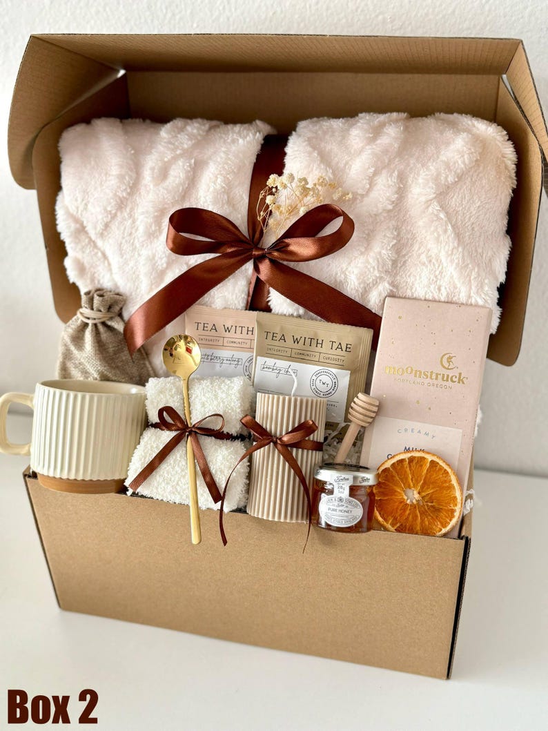 May include: A gift box filled with self-care items. It includes a fluffy white blanket, a mug, tea packets, a candle, honey, a wooden honey dipper, a bar of chocolate, and a dried orange slice. The box is tied with a brown ribbon.