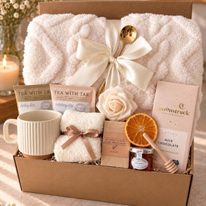 May include: A gift box containing a cream-colored cable knit blanket tied with a satin ribbon and gold spoon, a ribbed mug, two Tea With Tae tea bags, a rose, a small folded cloth, oatmeal soap, a dried orange slice, honey, and a Moonstruck chocolate bar.