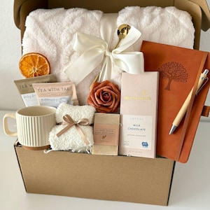 May include: A gift box filled with self-care items. Includes a cream-colored plush blanket tied with a bow, a mug, tea packets, a bar of soap, chocolate, a notebook, and a pen. A dried orange slice and a rose are also included. The box is labeled "Box 5".