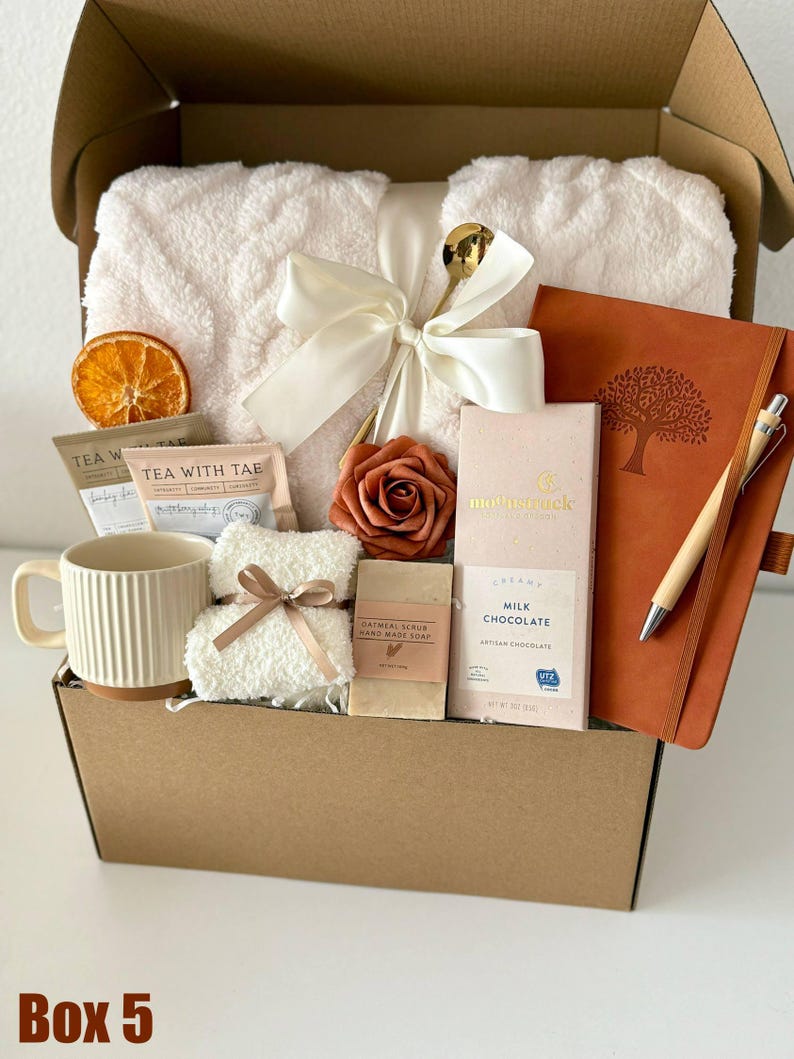 May include: A gift box filled with self-care items. Includes a cream-colored plush blanket tied with a bow, a mug, tea packets, a bar of soap, chocolate, a notebook, and a pen. A dried orange slice and a rose are also included. The box is labeled "Box 5".
