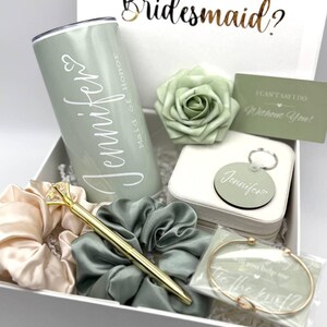 May include: A white gift box with a green and white bridesmaid proposal gift set. The box includes a tumbler, a green rose, a keychain, a bracelet, hair ties, and a pen. The text on the box reads "Will you be my bridesmaid?" and the tumbler says "Maid of Honor Jennifer".