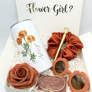May include: A gift box with the text "WILL YOU BE MY Flower Girl?" contains a white tumbler with orange flower graphics, a rust-colored scrunchie, sunglasses, a rose, and a bracelet. The tumbler is personalized with the name "Ashley".