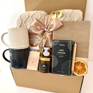 May include: A gift box filled with self-care items. Includes two mugs, a blanket, honey, soap, hot cocoa, a pen, and a dried orange slice. The box is brown with the text "Box 15" at the bottom.