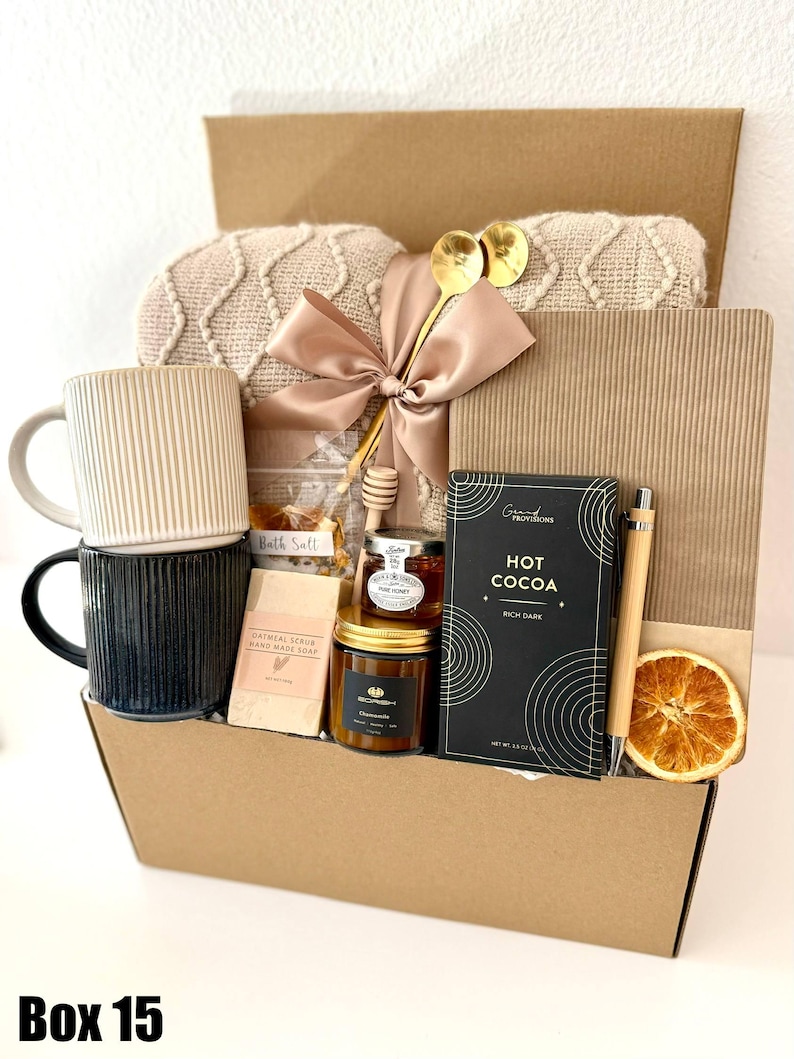 May include: A gift box filled with self-care items. Includes two mugs, a blanket, honey, soap, hot cocoa, a pen, and a dried orange slice. The box is brown with the text "Box 15" at the bottom.