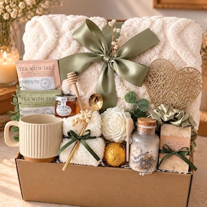 May include: A gift box containing a cream-colored textured throw blanket with a sage green ribbon, a ribbed ceramic mug, two Tea With Tae packets, a jar of honey, a honey dipper, a gold spoon, a white flower, a gold sphere, and oatmeal soap.