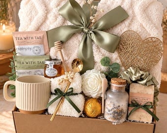 May include: A gift box containing a cream-colored textured throw blanket with a sage green ribbon, a ribbed ceramic mug, two Tea With Tae packets, a jar of honey, a honey dipper, a gold spoon, a white flower, a gold sphere, and oatmeal soap.
