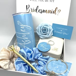 May include: A white gift box filled with items for a bridesmaid proposal. The box includes a blue tumbler with the name "Ashley" on it, a blue satin scrunchie, a gold pen, a white card with the text "I can't say I do without you!" and a blue rose.