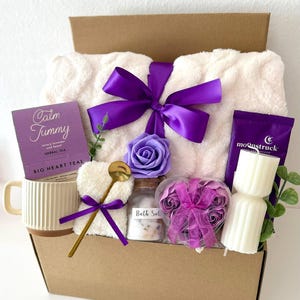May include: A gift box filled with self-care items. It includes a white plush blanket tied with a purple bow, a mug, tea, bath salts, a heart-shaped washcloth, a spoon, candles, and decorative flowers. The box is brown with the text "Box 10" at the bottom.