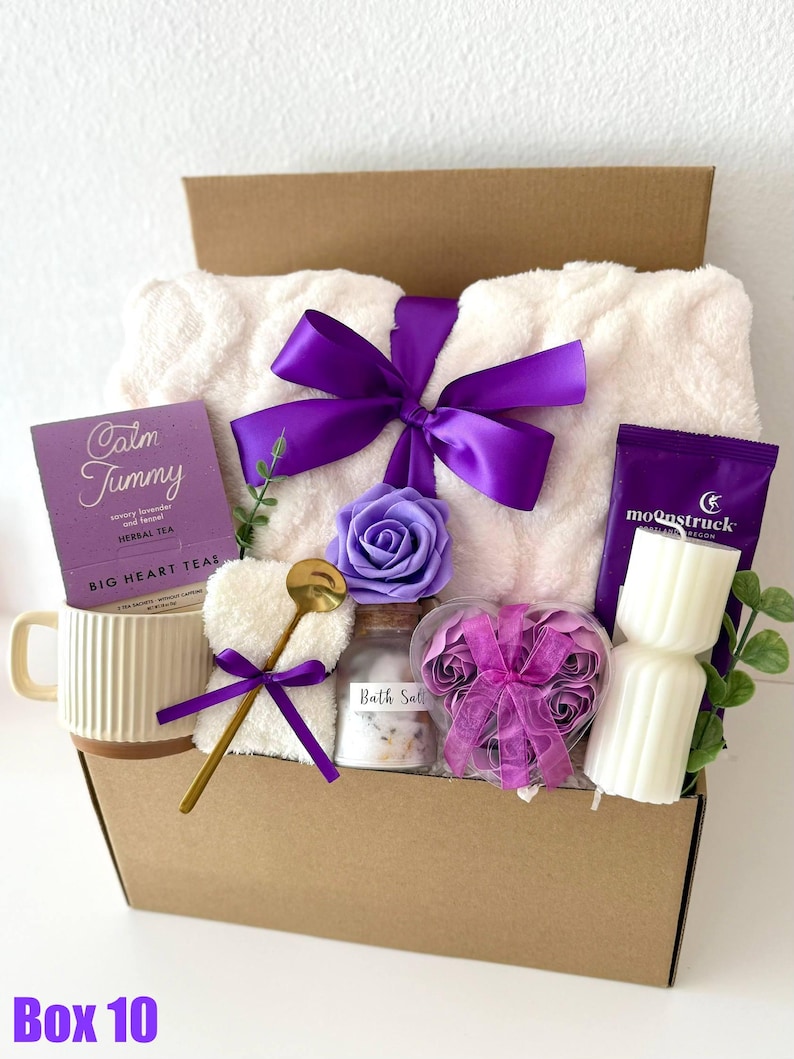 May include: A gift box filled with self-care items. It includes a white plush blanket tied with a purple bow, a mug, tea, bath salts, a heart-shaped washcloth, a spoon, candles, and decorative flowers. The box is brown with the text "Box 10" at the bottom.
