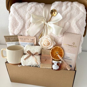 May include: A gift box filled with a cream-colored plush blanket tied with a white ribbon, a mug, tea packets, soap, chocolate, honey, and a decorative rose. The box is brown.