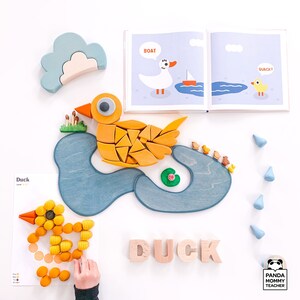 May include: A wooden toy duck puzzle with a blue pond and lily pad. The puzzle pieces are yellow and orange. The word "Duck" is spelled out in wooden letters. The image also includes a book with illustrations of a duck and a boat.