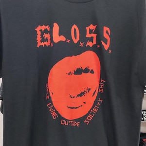 Puede incluir: Camiseta negra con un gráfico rojo. El gráfico incluye la palabra "GLOSS" en una fuente estilizada sobre un círculo rojo con un diseño similar a una cara. Debajo del círculo, el texto dice "SOCIETY'S SHIT" y "LIVING OUTSIDE".