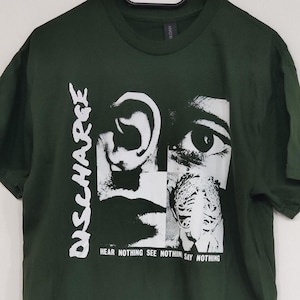 Può includere: Una t-shirt a maniche corte color verde bosco scuro con una stampa grafica bianca. Il design include il testo DISCHARGE in verticale e la frase HEAR NOTHING SEE NOTHING SAY NOTHING in orizzontale, insieme a immagini in bianco e nero di un occhio e un orecchio.