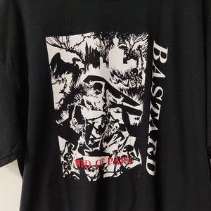 May include: Black t-shirt with a white and red graphic design. The graphic features a detailed illustration with the word "BASTARD" in white on the right side and "WILD OF PAIN" in red at the bottom. The shirt has a crew neck and short sleeves.