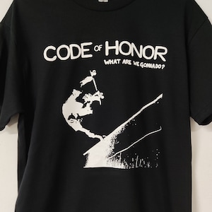Può includere: T-shirt nero con le scritte bianche "CODE OF HONOR" e "WHAT ARE WE GONNA DO?". Sotto il testo c'è una grafica bianca di una persona che cade. La maglietta è appesa a una gruccia nera.