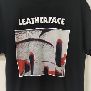 Op de afbeelding: Zwart T-shirt met een witte afbeelding van een band genaamd "Leatherface". De afbeelding toont een close-up van een groep paddenstoelen met rode en witte accenten.