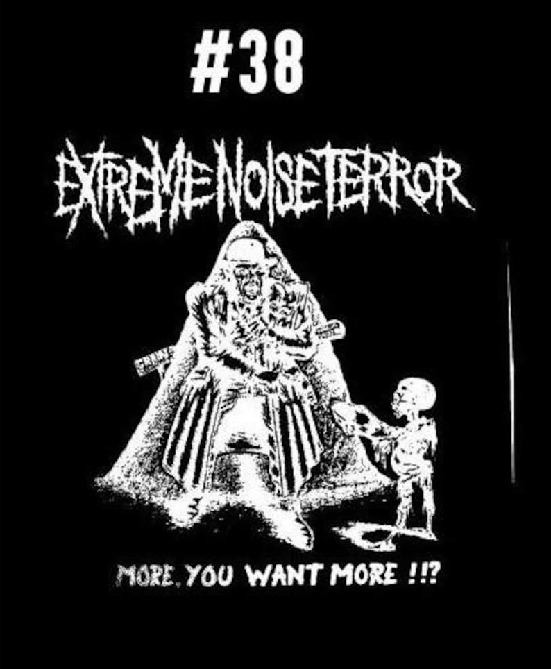 Puede incluir: Ilustraci&oacute;n en blanco y negro de un logotipo de banda, "Extreme Noise Terror", con el n&uacute;mero 38. La imagen presenta una figura con una capucha que sostiene un arma, y una figura m&aacute;s peque&ntilde;a de pie a la derecha. El texto "More, you want more!!" est&aacute; en la parte inferior de la imagen.