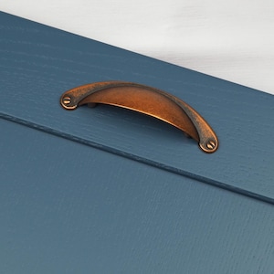 May include: A copper cabinet door pull with a rounded, arched shape. The pull is mounted on a blue painted wooden door.