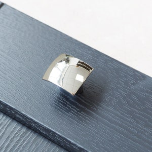 May include: A silver cabinet knob with a rounded, square shape. The knob is mounted on a dark grey cabinet door.