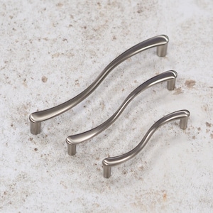 May include: Three silver-toned cabinet pulls with a wavy design. The pulls are arranged in order of size, with the largest pull at the top and the smallest pull at the bottom.