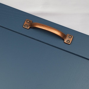 Op de afbeelding: Een koperen kastdeurgreep met een afgeronde rechthoekige vorm en een geborstelde afwerking. De greep is gemonteerd op een donkerblauwe geverfde deur.