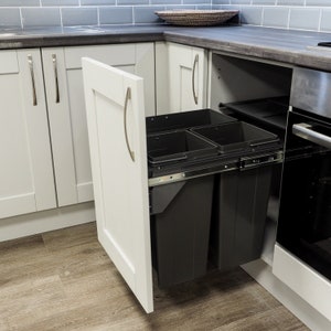 Puede incluir: Un mueble de cocina blanco con dos cubos de basura negros extraíbles. La puerta está abierta, revelando los cubos. La encimera es gris oscuro y el mueble tiene un tirador plateado.