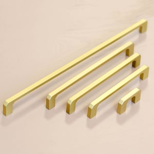 Könnte beinhalten: Fünf goldene Schrankgriffe in verschiedenen Größen. Die Griffe sind alle rechteckig mit abgerundeten Kanten und einem schlichten Design.